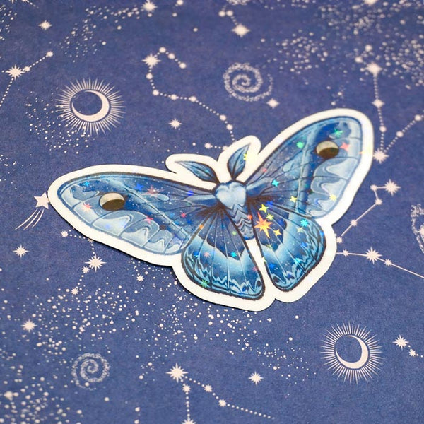 a waxing crescent moth holographic sticker on top of a blue starry paper from atelier persephone