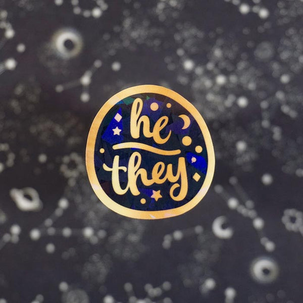 gold and holographic pronoun sticker of he they on a dark background from the shop atelier persephone
