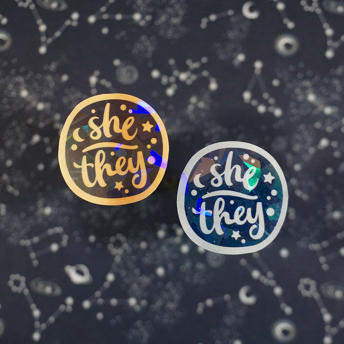 two stickers of she they on a black backgrpund from a lgbtq friendly business